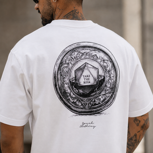 A man viewed from behind wearing a white t-shirt with a large black graphic print on the back. The graphic features a circular ornate frame surrounding a d20 die, with the text 'TAKE THE RISK' in the center.