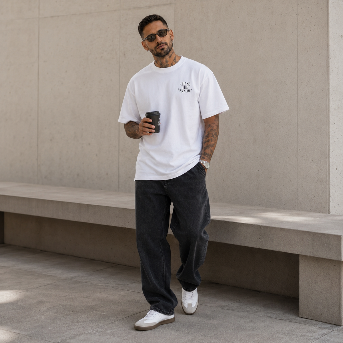 A man wearing a white crewneck t-shirt with a small black graphic on the left chest, paired with dark gray baggy jeans and white sneakers, standing in front of a concrete wall and holding a black coffee cup.