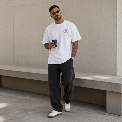 A man wearing a white crewneck t-shirt with a small black graphic on the left chest, paired with dark gray baggy jeans and white sneakers, standing in front of a concrete wall and holding a black coffee cup.