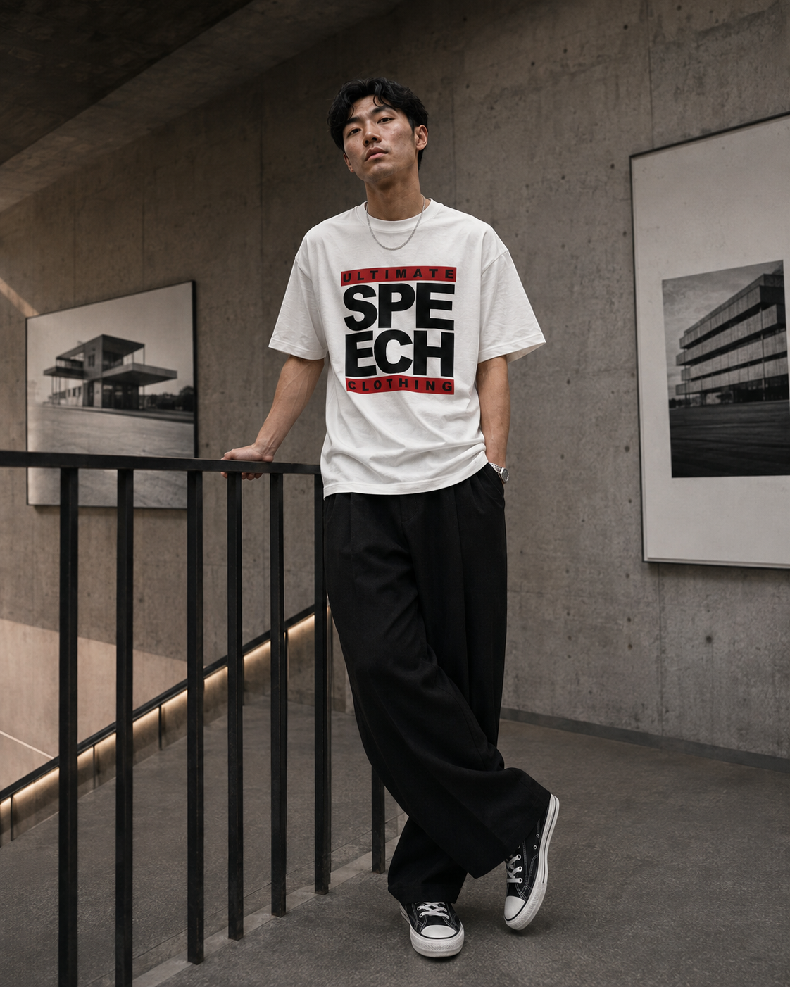 A man wearing a white graphic t-shirt with 'SPEECH DESIGN' in bold black and red letters, paired with black wide-leg pants and black sneakers, leaning against a black railing in a concrete hallway.