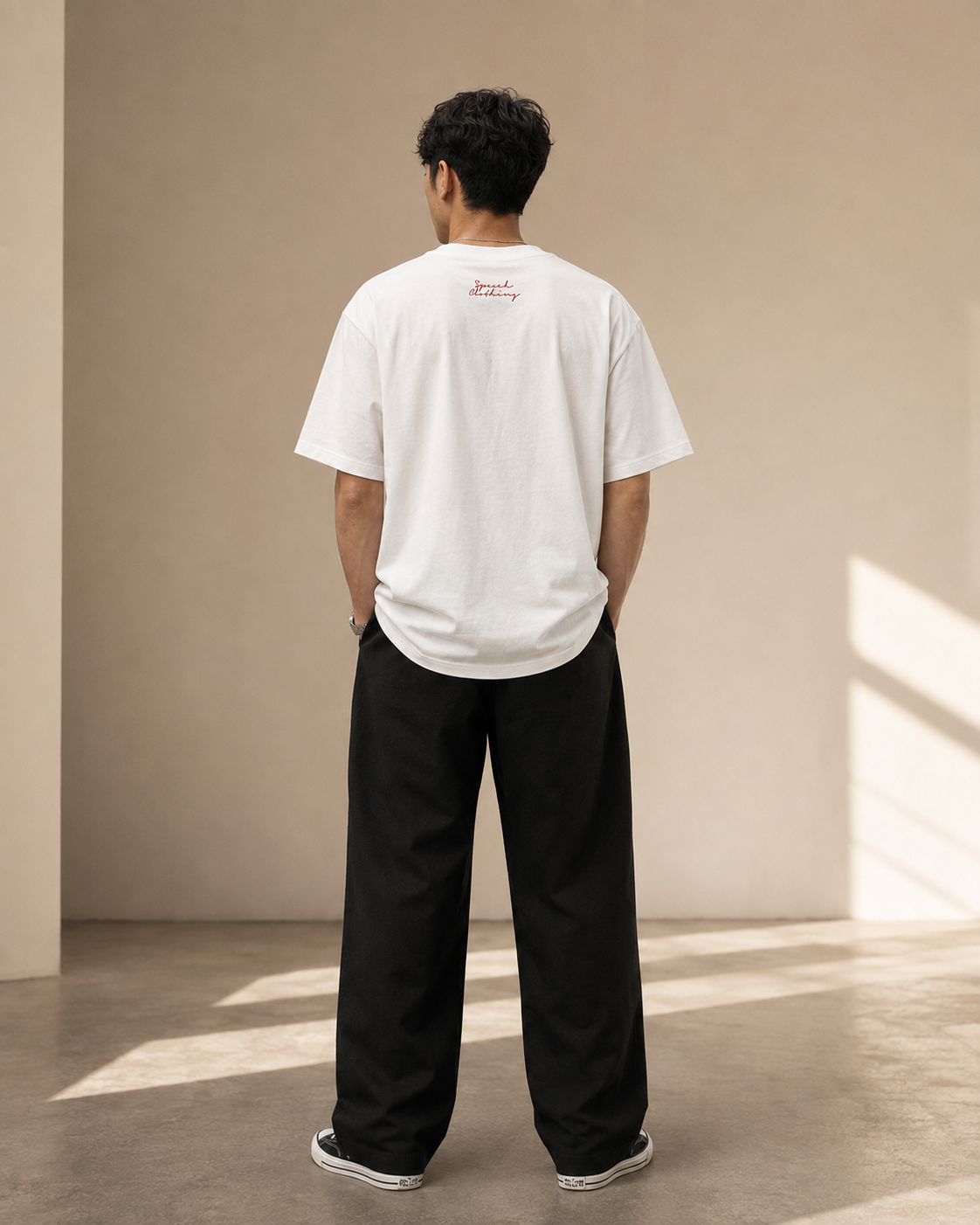 A man viewed from behind wearing a white crewneck t-shirt with a small red script logo on the upper back, paired with black wide-leg pants and white sneakers, standing in a minimalist beige room with soft window light.