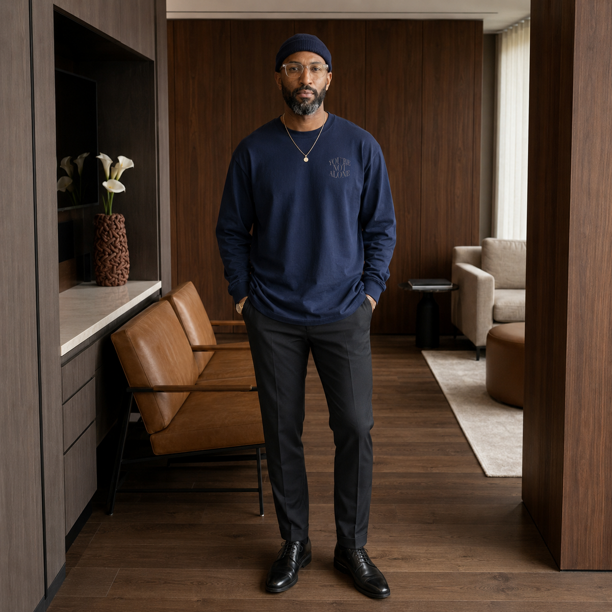 A man stands in a modern interior wearing a navy crewneck sweatshirt with a subtle embroidered logo on the left chest, dark trousers, black dress shoes, and a navy beanie.