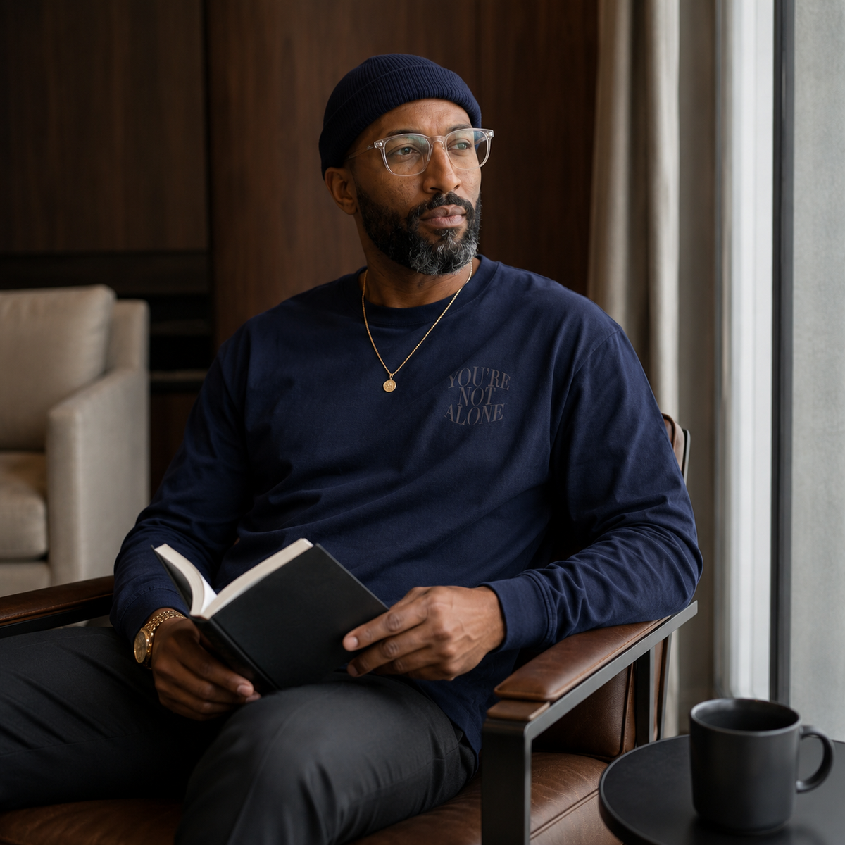 A man wearing a navy blue long-sleeve crewneck sweatshirt with 'YOU'RE ALONE' printed on the chest, sitting in a leather chair and holding an open book.