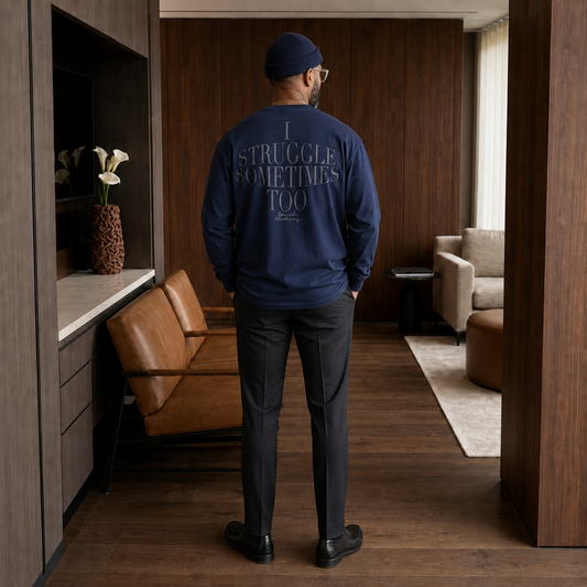 A man stands with his back to the camera in a modern living room, wearing a navy blue crewneck sweatshirt with the text 'I STRUGGLE SOMETIMES TOO' printed in light gray on the back, paired with dark gray trousers and a navy beanie.