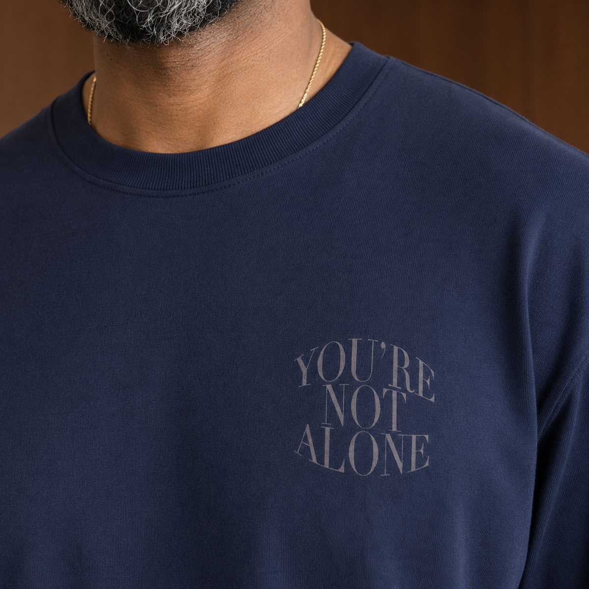 A close-up of a person wearing a navy blue crewneck sweatshirt with the words 'YOU'RE NOT ALONE' printed in a muted grayish tone on the left chest. The person has a gold chain necklace, and the background is a warm brown wood panel.