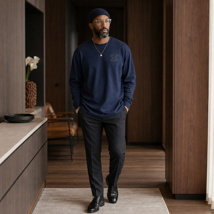 A man wearing a navy blue crewneck sweatshirt with a subtle embroidered logo on the left chest, black trousers, and black leather shoes, walking in a modern hallway with dark wood paneling.
