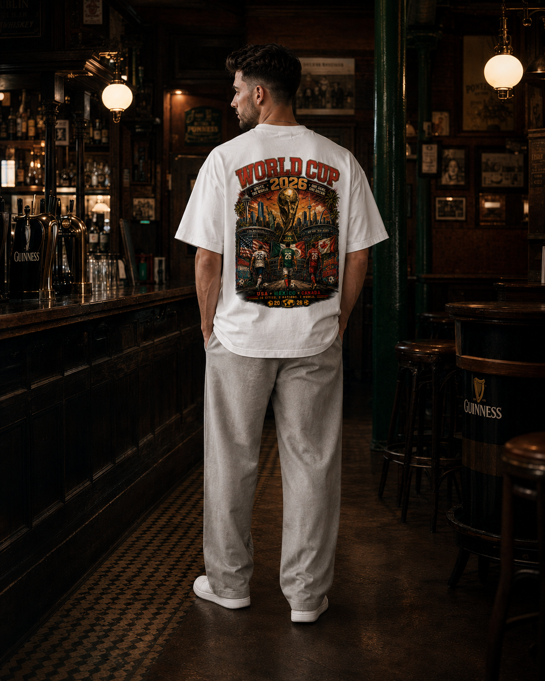 A man stands with his back to the camera in a dimly lit pub, wearing a white t-shirt with a large colorful graphic print on the back that reads 'WORLD CUP 2026' and 'MEXICO'.